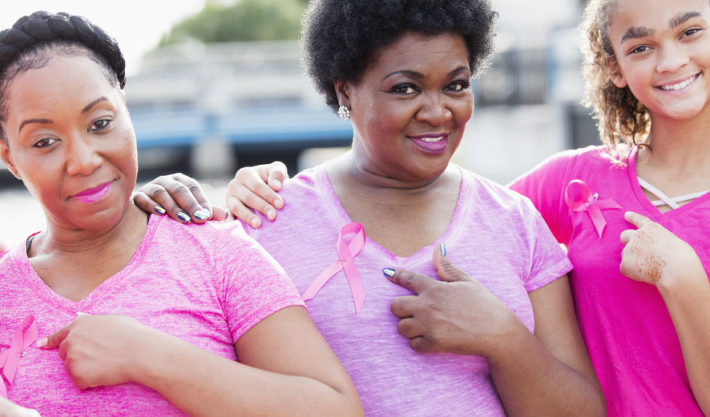 A group of three multi-ethnic women of mixed ages standing together at a breast cancer awareness event, wearing pink, pointing to the ribbons pinned to their shirts. The youngest, on the right, is a mixed race African-American and Caucasian teenage girl. The mature African-American woman in the middle is in her 50s and her friend is in her 30s.